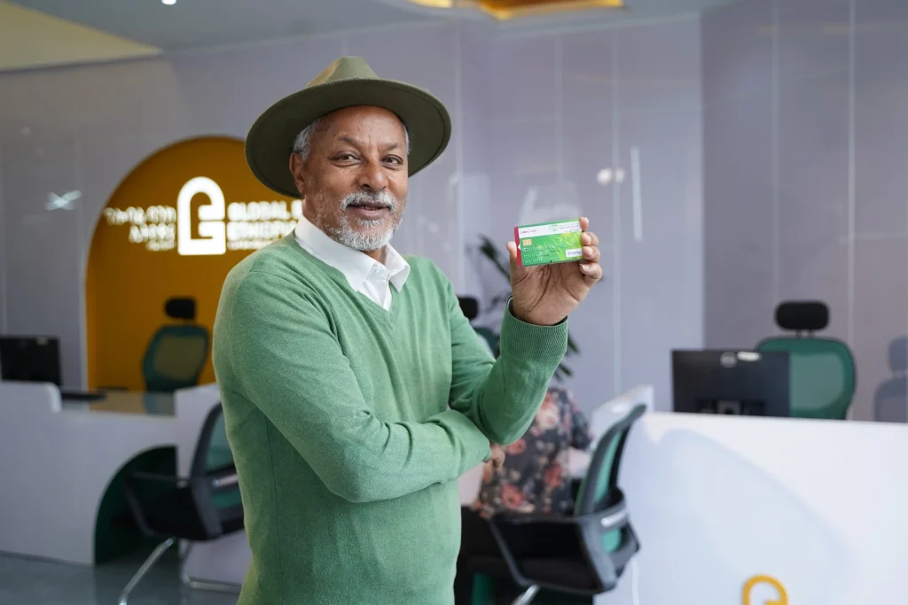 An elderly man in a green sweater and hat proudly holding a Global bank ATM card inside a Global Bank branch and taking a picture.